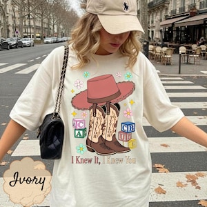 May include: An ivory t-shirt featuring a graphic of a pink cowboy hat, two brown cowboy boots labeled Taylor, and colorful alphabet blocks. The design includes small flowers and the text I Knew It, I Knew You. A black shoulder bag is also visible.