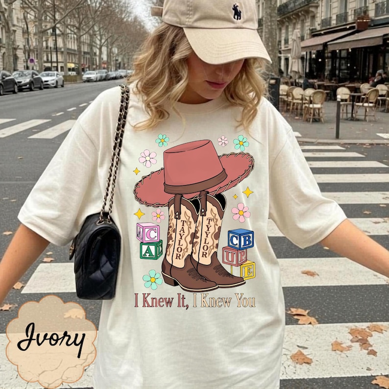 May include: An ivory t-shirt featuring a graphic of a pink cowboy hat, two brown cowboy boots labeled Taylor, and colorful alphabet blocks. The design includes small flowers and the text I Knew It, I Knew You. A black shoulder bag is also visible.