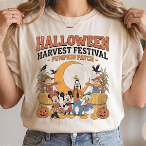 May include: An ivory t-shirt featuring a graphic print with the text HALLOWEEN HARVEST FESTIVAL PUMPKIN PATCH. The design includes Mickey Mouse, Minnie Mouse, Goofy, and Donald Duck surrounded by scarecrows, hay bales, pumpkins, and a crescent moon.