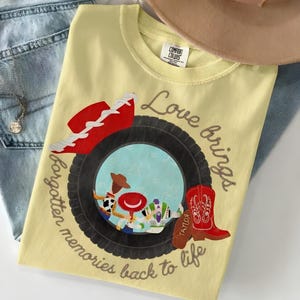 May include: A pale yellow t-shirt featuring a graphic of a black tire framing a scene with three cartoon characters. The design includes a red cowboy hat, a red cowboy boot with the name Taylor, and the text Love brings forgotten memories back to life.