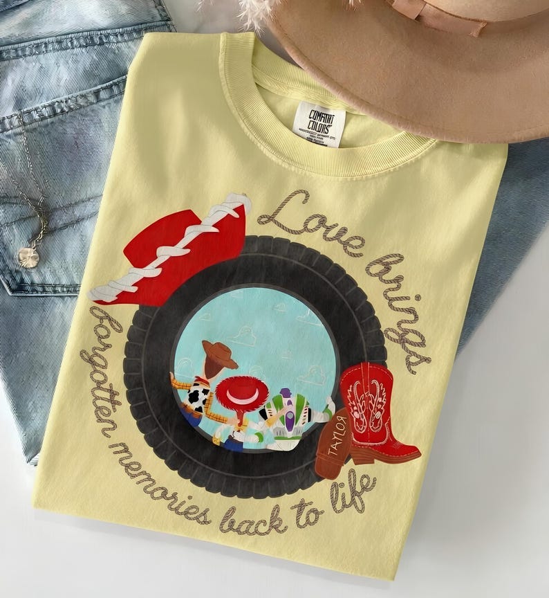 May include: A pale yellow t-shirt featuring a graphic of a black tire framing a scene with three cartoon characters. The design includes a red cowboy hat, a red cowboy boot with the name Taylor, and the text Love brings forgotten memories back to life.