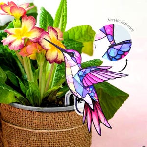 May include: A colorful acrylic hummingbird garden stake. The hummingbird has a stained-glass design with pink, purple, blue, and white hues. The stake is placed in a burlap-wrapped flower pot with yellow and pink flowers.