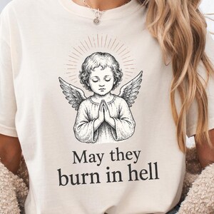 以下が含まれることがあります： クリーム色の半袖Tシャツ。手を合わせ翼を広げた天使のイラストが黒いインク調で描かれ、その周囲には光輪が添えられている。グラフィックの下には「May they burn in hell」というテキストが黒色でプリントされている。