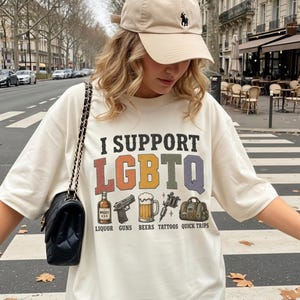May include: A beige baseball cap with a small dark logo and an oversized cream t-shirt featuring the text I SUPPORT LGBTQ with colorful block letters and illustrations of liquor, guns, beer, tattoos, and a duffel bag labeled quick trips.