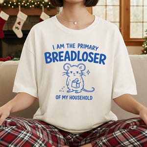Op de afbeelding: Een gebroken wit T-shirt met korte mouwen en een blauwe afbeelding van een muis die een brood vasthoudt. De tekst luidt: I AM THE PRIMARY BREADLOSER OF MY HOUSEHOLD. Het shirt wordt gecombineerd met een rood-zwarte pyjamabroek met ruitjes.
