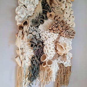 Può includere: Un arazzo da parete strutturato con una varietà di elementi all'uncinetto e tessuti in tonalità di bianco, beige, grigio e marrone. L'opera presenta forme tridimensionali che ricordano coralli e altre forme organiche, montate su un'asta di legno.