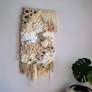 May include: A textured wall hanging with a variety of off-white, beige, and tan yarns. The woven artwork features a mix of looped, fringed, and knotted textures, creating a three-dimensional effect. It is mounted on a natural wooden dowel.