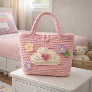 May include: A pink crochet tote bag with a rectangular shape and two handles. The bag is decorated with a cloud and heart applique, along with three flower appliques in yellow, purple, and green. The bag is sitting on a white table.