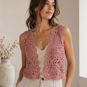 Puede incluir: Un chaleco de ganchillo en color rosa empolvado con un patrón de motivos florales repetitivos y un borde festoneado a lo largo de la abertura frontal y el dobladillo. La prenda sin mangas se lleva sobre una camiseta beige y pantalones claros.
