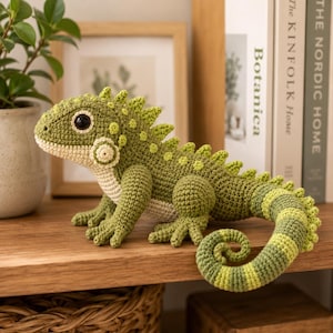 Può includere: Un iguana all'uncinetto fatto a mano, nei toni del verde e del crema, con squame dettagliate e una coda arricciata. Il rettile è posizionato su una mensola di legno, mettendo in risalto il suo corpo strutturato e i grandi occhi scuri. Un oggetto decorativo unico.