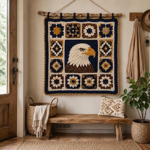 May include: A handcrafted, square wall hanging featuring a detailed crochet design of an eagle's head. The artwork is framed by a navy blue border and adorned with floral and geometric patterns in shades of brown, cream, and gold. The piece is suspended from a wooden rod.