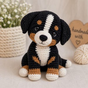 Puede incluir: Un cachorro de perro de montaña bernés hecho a mano con ganchillo. El peluche presenta hilo negro, blanco y marrón, con una raya blanca en la cara. El cachorro tiene grandes ojos negros y está sentado sobre una superficie clara.