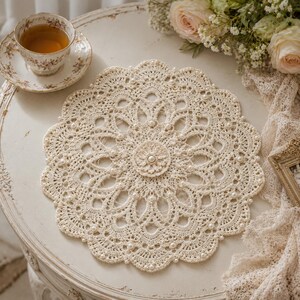Könnte beinhalten: Cremefarbene, gehäkelte Tischdecke mit gewelltem Rand und Perlenakzenten. Die Tischdecke hat ein zentrales Blumendesign und liegt auf einem weißen, antiken Tisch. Eine Teetasse und Untertasse sind im Hintergrund zu sehen.