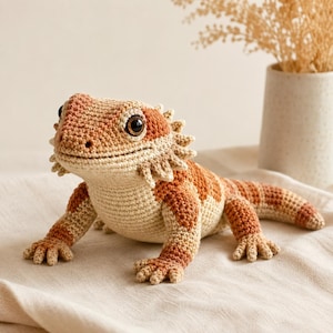 May include: A crocheted bearded dragon in shades of beige and orange. The handmade reptile features detailed scales, a spiky frill, and large, expressive eyes. The plush toy is posed on a neutral surface, showcasing its intricate design.