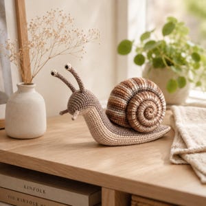 Puede incluir: Una figura de caracol tejida a ganchillo con un cuerpo de color beige claro y una concha en espiral con bandas alternas de hilo marrón, tostado y crema. El caracol tiene dos tallos largos con ojos oscuros y dos tentáculos cortos.
