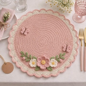 Könnte beinhalten: Ein handgefertigter, runder, rosa gehäkelter Platzdeckchen mit einem gewellten Rand. Der Platzdeckchen hat einen cremefarbenen Rand und ist mit gehäkelten Schmetterlingen, Blumen und Blättern verziert. Die Blumen sind rosa, weiß und gelb.