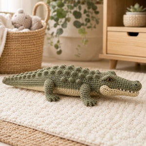 May include: A handmade crochet crocodile toy featuring a textured olive green back with raised bumps, a light cream-colored underbelly, and a long tail. The creature has small dark eyes and a wide snout with visible white stitched teeth.
