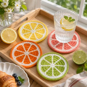 Può includere: Un vassoio di legno contiene quattro sottobicchieri all'uncinetto a forma di fette di agrumi: limone, arancia, pompelmo e lime. Un bicchiere d'acqua con limone e menta è appoggiato sul sottobicchiere di pompelmo.