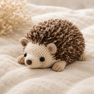 May include: A handmade crochet hedgehog toy. The hedgehog has a light beige face, ears, and paws, with a brown snout and black eyes. Its back is covered in brown and beige yarn, giving it a textured appearance. The toy is lying on a cream-colored surface.