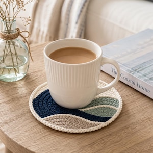 Op de afbeelding: Een witte keramische mok gevuld met een lichtbruine drank staat op een handgemaakte gehaakte onderzetter. De onderzetter heeft een golfontwerp in marineblauw, lichtgroen en beige, met een witte rand. Een vaas met gedroogde bloemen en een boek staan ook op de houten tafel.