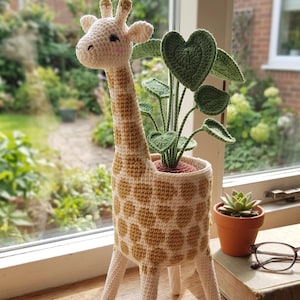 May include: A handcrafted crochet giraffe planter with a tan and white spotted pattern. The giraffe holds a green leafy plant in its head. A small terracotta pot with a succulent and a pair of glasses sit nearby on a wooden windowsill.
