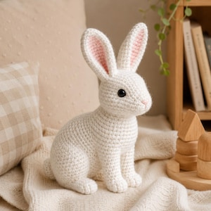 May include: A white crocheted rabbit stuffed animal with long ears featuring light pink inner detailing. The rabbit has black plastic eyes and a small pink stitched nose. It is crafted with a visible yarn texture and sits in an upright position.