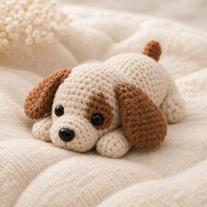 May include: A small, handmade crochet dog toy crafted from cream-colored yarn with brown patches on the ears and around one eye. The plush figure features two shiny black plastic eyes and a small black plastic nose, with a short tail.