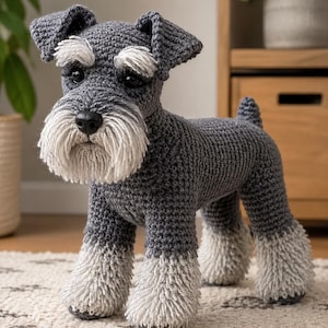 May include: A crocheted Schnauzer dog figurine featuring a dark grey body with light grey textured fur on the muzzle, eyebrows, and lower legs. The toy has black plastic eyes and a black nose, standing on four legs with a short upright tail.