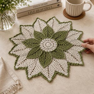 May include: A handmade crochet doily in a floral design. The doily features a central green flower surrounded by white petals with green edging. The doily is approximately 30 cm in diameter, suitable for a table centerpiece.