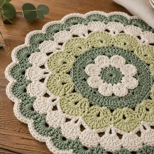 May include: A handmade, circular crochet doily in shades of green and cream. The doily features a floral design in the center and a scalloped edge. The yarn appears to be cotton, and the doily is placed on a wooden surface.