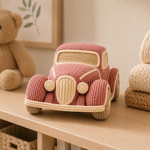 May include: A crocheted toy car in dusty rose and cream yarn. The vehicle features a rounded body, a prominent front grille, two circular headlights, and four wheels, all crafted with a textured stitch pattern in a two-tone color scheme.