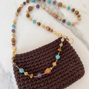 May include: A brown crocheted handbag with a beaded strap. The strap features a variety of beads in colors like turquoise, amethyst, tiger's eye, and wood. Gold-colored clasps connect the strap to the bag.