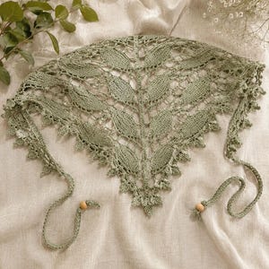 Könnte beinhalten: Ein salbeigrünes, dreieckiges Kopftuch mit einem komplizierten Strickmuster aus Blättern und Lochstickerei. Die Kanten sind mit einer zarten Bogenkante versehen, und die beiden dünnen Bänder sind jeweils mit einer kleinen Holzperle verziert.