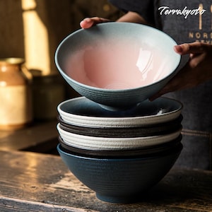 May include: A stack of ceramic bowls in various colors. The top bowl is held up, revealing a pink interior that fades to white. The exterior is a light blue. Other bowls are dark blue, black, and white. The bowls have a textured, ribbed design.