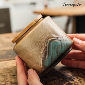 Puede incluir: Un recipiente cerámico cuadrado con tapa de madera. El recipiente es de color beige con un diseño de montaña turquesa en una esquina. El texto "Terrakyto" está en la esquina superior derecha.