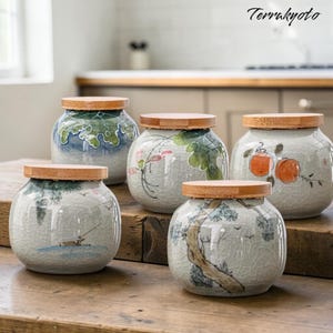 Könnte beinhalten: Fünf Keramikgläser mit Holzdeckeln. Jedes Glas hat ein einzigartiges handgemaltes Design in Blau-, Grün- und Orangetönen. Die Gläser stehen auf einer Holzoberfläche und zeigen ihre dekorativen und funktionalen Eigenschaften.