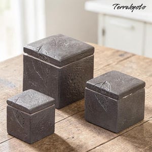 Könnte beinhalten: Drei dunkelgraue quadratische Boxen mit strukturierten Oberflächen und passenden Deckeln sind auf einem Holztisch angeordnet. Die Boxen variieren in der Größe, die größte in der Mitte und die kleinste links. Die Boxen haben ein rustikales, steinartiges Aussehen.