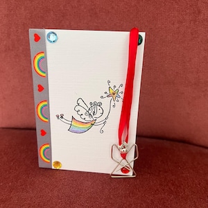 Op de afbeelding: Handgemaakte wenskaart met een grillige engelillustratie, een regenboogvleugel en een ster. De kaart heeft een grijze rug met regenboog- en hartaccenten. Een zilveren engelbedel met een rode kraal hangt aan een rood lint.