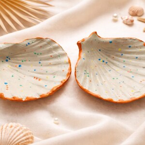 Peut inclure: Deux vide-poches en forme de coquillage, intérieurs blancs et bords orange. Les plats sont décorés d'éclaboussures de peinture colorées. Le fond comprend des perles, des coquillages et une étoile de mer sur un tissu crème.