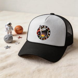 May include: A black and white trucker hat with a mesh back and a curved brim. The front features a circular patch with the text "PROJECT HAIL MARY" and a rocket design. Miniature astronaut, planet, and star figurines are in the background.