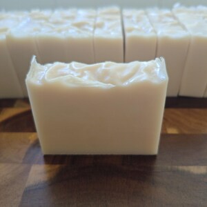 May include: A close-up of a light beige bar of soap with a textured top, in front of a row of similar bars. The soap has a creamy appearance and is on a dark wooden surface.