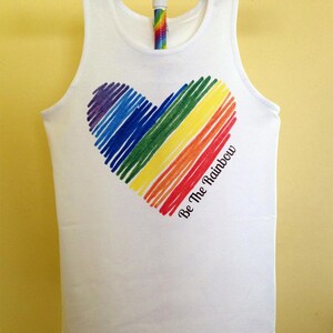 May include: White tank top with a rainbow heart design. The heart is composed of colorful, diagonal lines in red, orange, yellow, green, blue, and purple. The text "Be The Rainbow" is printed on the lower right side of the heart.