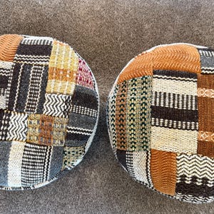 May include: Two round, patchwork poufs with a variety of woven fabric patterns in shades of brown, beige, black, gray, and green. The poufs are placed side by side on a neutral-colored surface.