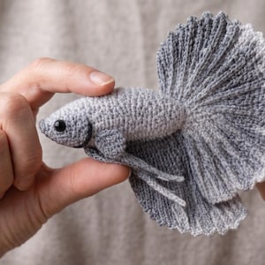 Puede incluir: Un pez Betta de ganchillo gris hecho a mano, con una gran cola en forma de abanico y aletas detalladas. El pez tiene un ojo oscuro y se sostiene entre dos manos. El pez mide aproximadamente 10 cm de largo.