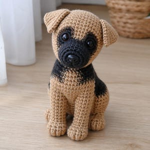 Könnte beinhalten: Gehäkelte Welpenfigur in Beige und Schwarz, die auf einer Holzoberfläche sitzt. Der handgefertigte Hund hat eine schwarze Schnauze und Augen, mit beigen Ohren und Körper. Ein bezaubernder Dekorationsartikel.
