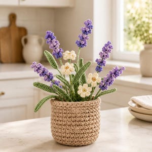 Puede incluir: Un arreglo floral de ganchillo con lavanda y flores tipo margarita en una maceta beige. La lavanda tiene capullos morados y azules, mientras que las margaritas son de color crema. La maceta es de color beige claro y está sobre una encimera blanca.