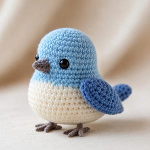 Op de afbeelding: Een handgemaakte gehaakte vogel in licht- en donkerblauwe tinten en crème. De vogel heeft een afgerond lichaam, donkere ogen en een grijze snavel en poten. De vleugels en staart zijn donkerblauw. Een charmant decoratief item.