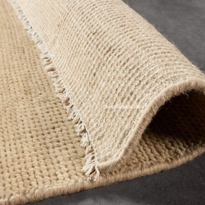 May include: A beige area rug with a thick, textured, hand-woven loop pile construction. The rug features a folded corner revealing the underside and a short, knotted fringe along the edge. The text CreamOushakStore is visible on the surface.