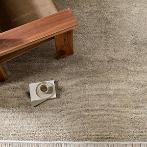 May include: A rectangular area rug with a textured, beige, woven surface and a short white fringe along the bottom edge. The rug features a consistent, dense loop pile construction that covers the entire surface area in a neutral, earthy tone.