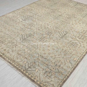 Op de afbeelding: Een rechthoekig vloerkleed met een gedempt kleurenpalet. Het vloerkleed heeft een herhalend geometrisch patroon in tinten beige, grijs en lichtblauw. Het ontwerp bevat gestileerde bloem- en bladmotieven. Het vloerkleed heeft een gefranjerde rand.